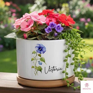 May include: White ceramic flower pot with a floral design and the name "Victoria" in script. The pot is filled with pink, red, and purple flowers, and green trailing plants. It sits on a wooden saucer.