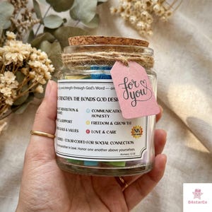 May include: A glass jar with a cork lid, tied with twine, and a pink tag that says "for you." The jar contains colourful paper strips and has text on the front. The text includes "STRENGTHEN THE BONDS GOD DESI" and "COMMUNICATION HONESTY".
