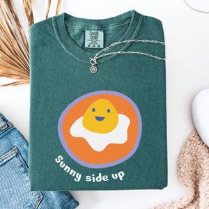 May include: A dark teal crewneck t-shirt featuring a graphic of a smiling fried egg inside an orange circle with a light purple border. The text "Sunny side up" is printed in white below the graphic. A silver chain necklace rests on the collar.