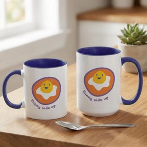 May include: Two white ceramic mugs with deep blue interiors and handles. Each mug features a graphic of a smiling fried egg inside an orange circle with the text Sunny side up printed below. A silver metal spoon lies horizontally in front.