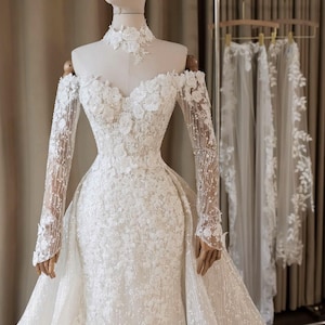 Op de afbeelding: Een witte vloerlange japon met lange transparante mouwen, een off-shoulder halslijn en een kanten choker. Het lijfje en de rok zijn versierd met ingewikkelde bloemenkantapplicaties, kralenwerk en een volumineuze afneembare overrok.