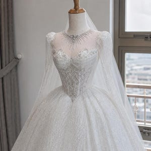 Op de afbeelding: Een witte, vloerlange baljurk met lange transparante mouwen, een hartvormig lijfje en een hoge illusie-halslijn. De jurk is rijkelijk versierd met zilveren pailletten, kralenwerk en kristallen, gecombineerd met een bijpassende transparante sluier.