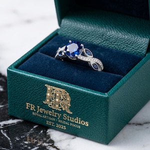 Puede incluir: Un anillo de compromiso plateado con una gran piedra preciosa azul redonda y piedras azules más pequeñas, engastadas en una banda decorativa. El anillo se muestra en una caja verde con el texto "FR Jewelry Studios" y "Royal Artisan - Bridal Focus EST. 2025".