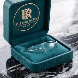 May include: A silver bracelet with a mermaid tail design and a pearl, displayed in an open teal jewellery box. The box has a velvet lining and the text "FR Jewelry Studios Royal Artisan Bridal Focus EST 2025".