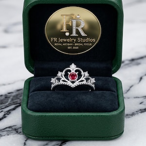 Puede incluir: Un anillo tipo tiara plateado con una piedra roja redonda central y pequeñas piedras claras engastadas en un diseño de corona. El anillo se presenta dentro de un estuche verde oscuro forrado de terciopelo con el logo dorado FR Jewelry Studios.