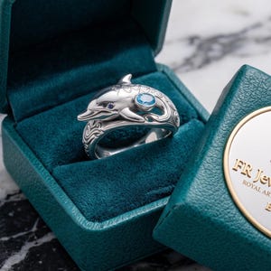 May include: A silver ring shaped like a dolphin with a small blue gemstone set into its body, resting inside a teal velvet-lined jewelry box. The box lid features a gold and silver emblem with the text FR Jewelry Studios ROYAL ARTISAN EST. 2025.