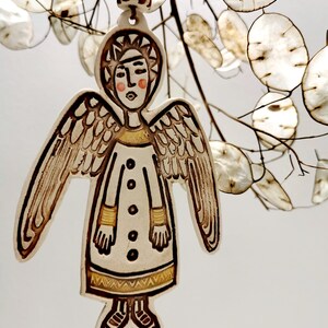 May include: A ceramic angel ornament with brown wings, a white dress, and a gold trim. The angel has a brown halo and a painted face with pink cheeks.