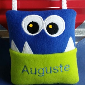 May include: A blue and green plush monster with white teeth and yellow eyes. The monster has a white cord loop at the top and a green pocket with the name "Auguste" embroidered on it.
