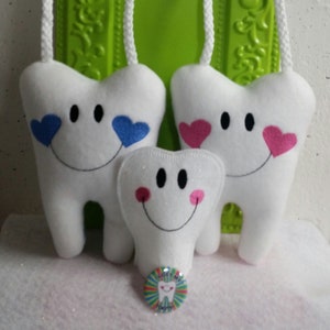 May include: Three white plush teeth with smiling faces and embroidered hearts. The two larger teeth have hanging loops. The smaller tooth has glitter and pink cheeks. A colorful button with the text "Lost My Tooth" is in the foreground.