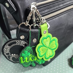 May include: Black leather bag with silver zippers and hardware. Attached are three keychains: a black circular keychain with a white embroidered design, a green clover keychain with the word "Lucky", and a bright green four-leaf clover keychain.