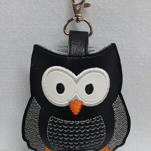 May include: Black faux leather owl-shaped hand sanitizer holder with a silver metal clip. The owl has white eyes, an orange beak, and a gray and black patterned body.