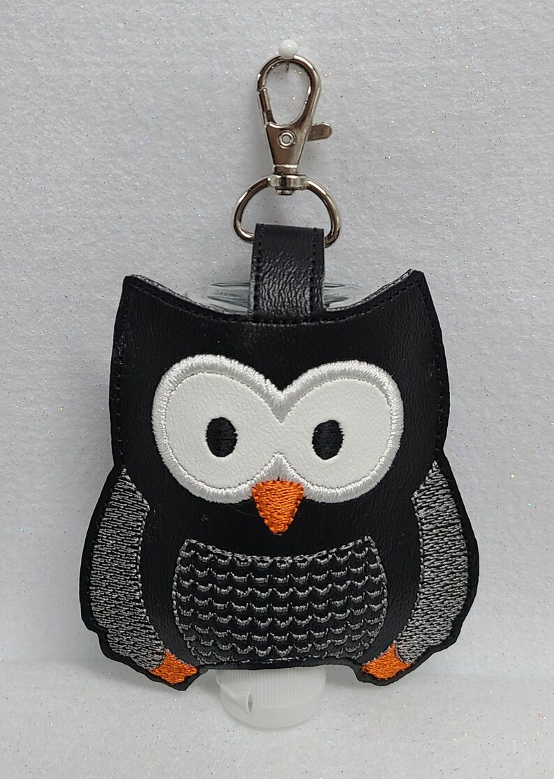 May include: Black faux leather owl-shaped hand sanitizer holder with a silver metal clip. The owl has white eyes, an orange beak, and a gray and black patterned body.