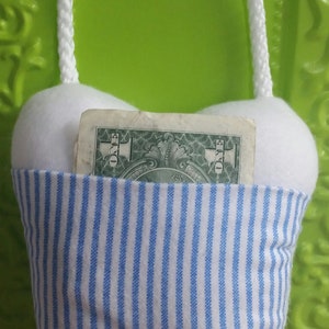 May include: A white tooth-shaped pillow with blue and white stripes. The pillow has a white rope handle and a dollar bill tucked inside the pocket.