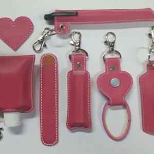 May include: Set of seven pink leather keychains with various shapes and sizes, including a heart, a rectangle, and a tube. Each keychain has a silver metal clasp and a white stitching detail.