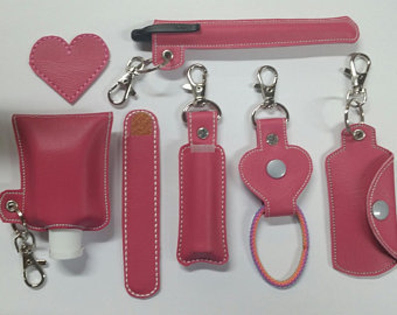 May include: Set of seven pink leather keychains with various shapes and sizes, including a heart, a rectangle, and a tube. Each keychain has a silver metal clasp and a white stitching detail.