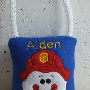 May include: Blue tooth pillow with a white tooth embroidered on it wearing a red firefighter hat with a yellow badge. The pillow has a white drawstring and the name Aiden is embroidered on it in yellow.