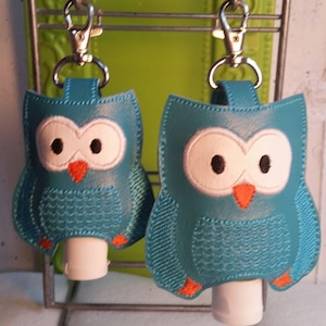 May include: Two teal blue owl-shaped hand sanitizer holders with orange beaks and white eyes. Each holder has a silver clip and a clear plastic bottle inside.