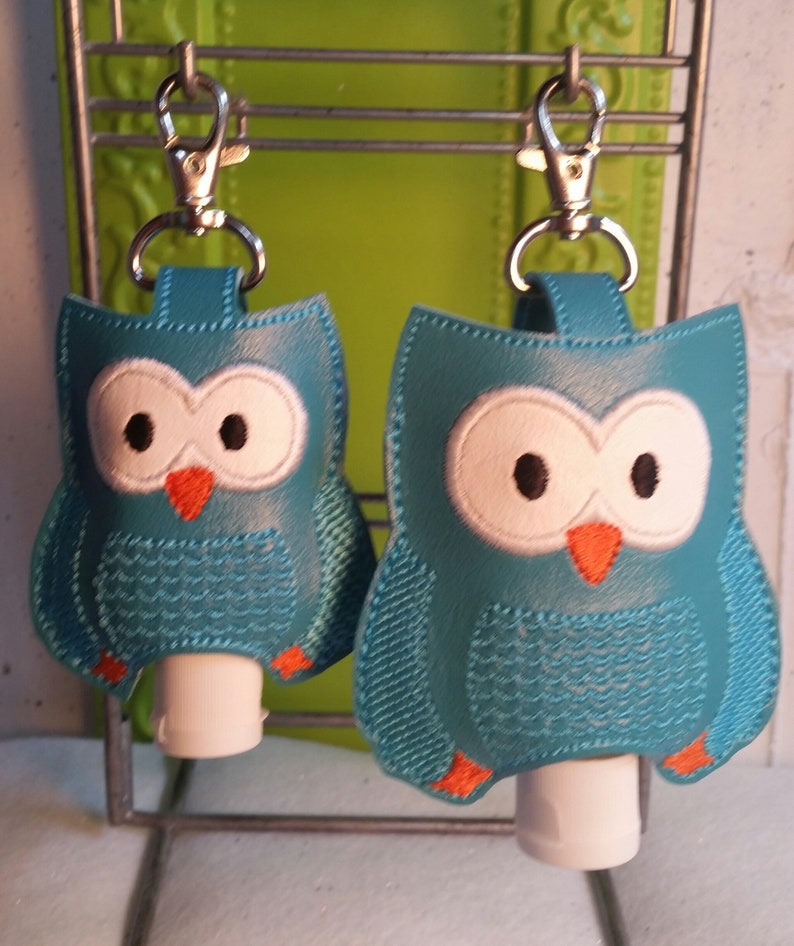 May include: Two teal blue owl-shaped hand sanitizer holders with orange beaks and white eyes. Each holder has a silver clip and a clear plastic bottle inside.