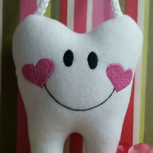 May include: A white plush tooth with a smiling face and pink hearts on the cheeks. The tooth is hanging from a white cord and has a round button with the text "I Lost My Tooth" on it.