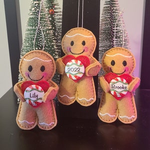 May include: Three felt gingerbread man ornaments with candy cane hearts. The hearts have the names "Lily", "Brooke", and "2022" written on them.