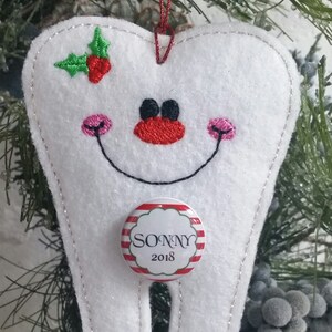 May include: White felt tooth ornament with a red and white striped candy cane button that says "SONNY 2018". The tooth has a smiling face with pink cheeks and black eyes. There is a green holly sprig with red berries on the top of the tooth.