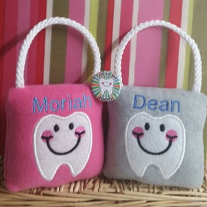 May include: Two personalized tooth fairy pillow bags, one pink with the name Moriah and one gray with the name Dean. Each bag features a white embroidered tooth with a smiling face.