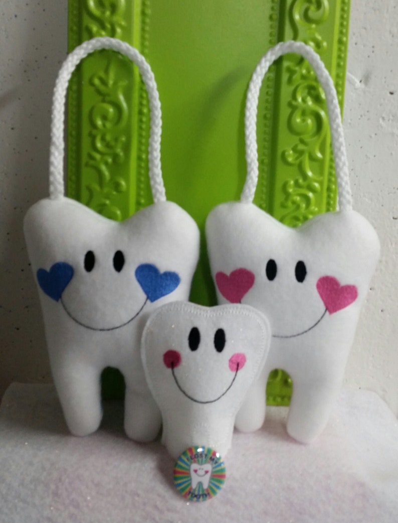 May include: Three white plush teeth with smiling faces and embroidered hearts. The two larger teeth have hanging loops. The smaller tooth has glitter and pink cheeks. A colorful button with the text "Lost My Tooth" is in the foreground.