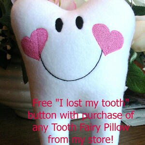 May include: White tooth-shaped pillow with pink embroidered hearts and a black embroidered smile. The text "Free "I lost my tooth" button with purchase of any Tooth Fairy Pillow from my store!" is written below the pillow.