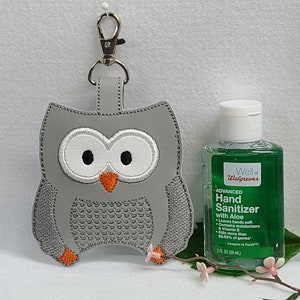 May include: Gray owl-shaped hand sanitizer holder with a silver keychain. The holder is made of felt and has a stitched owl design. A small bottle of hand sanitizer is next to the holder. The bottle is green and says "Well at Walgreens" and "Advanced Hand Sanitizer with Aloe" on the label.