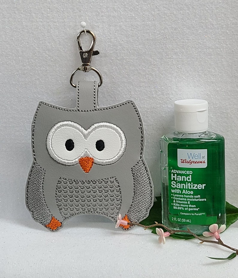 May include: Gray owl-shaped hand sanitizer holder with a silver keychain. The holder is made of felt and has a stitched owl design. A small bottle of hand sanitizer is next to the holder. The bottle is green and says "Well at Walgreens" and "Advanced Hand Sanitizer with Aloe" on the label.