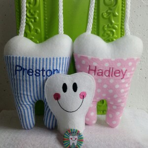 May include: Two plush tooth-shaped ornaments, one blue and white striped with the name "Preston" embroidered on it, and the other pink with white polka dots and the name "Hadley" embroidered on it. A smaller white plush tooth with a smiley face is in the center. A button with a smiling tooth and the words "I lost my tooth" is in the foreground.