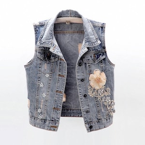 May include: A light blue denim vest with a distressed look. The vest features a collar, button closure, and decorative floral embellishments in cream and silver tones. The vest has two front pockets.