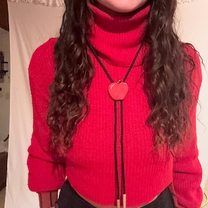 May include: A red turtleneck sweater with a matching apple-shaped bolo tie. The bolo tie has a black cord and gold-colored metal tips. The sweater has long sleeves and a ribbed texture. The outfit is completed with black pants.