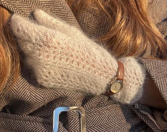May include: A pair of light gray, sheer, knitted mohair gloves with a delicate openwork pattern. The gloves are worn with a brown leather-strapped analog wristwatch and a brown and white herringbone patterned coat with a silver belt buckle.