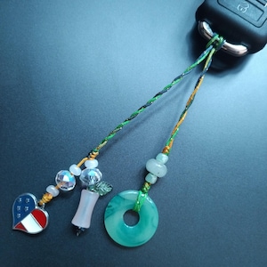 Puede incluir: Un llavero con abalorios decorativos. Los abalorios incluyen un corazón con un diseño rojo, blanco y azul, cuentas facetadas transparentes, un abalorio rosa y blanco y una rosquilla de jade verde. El llavero está sujeto a una llave de coche negra.