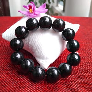 May include: A black beaded bracelet with large, round beads. The bracelet is displayed on a white square cushion, with a pink and yellow flower in the background. The beads have a glossy finish.