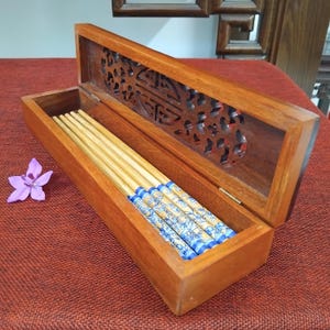 May include: Wooden chopstick box with a carved lid, containing several pairs of chopsticks. The chopsticks have blue and white floral designs on the ends. A purple flower rests beside the box. The box is made of a warm-toned wood.