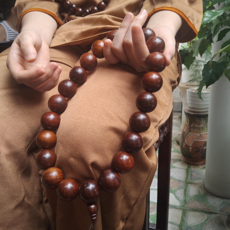 Op de afbeelding: Een lange streng van grote, gepolijste, donkerbruine houten gebedskralen. De kralen zijn bolvormig en van gelijke grootte, verbonden door een koord, en vormen een doorlopende lus met een kleinere kraal als accent aan de onderkant.