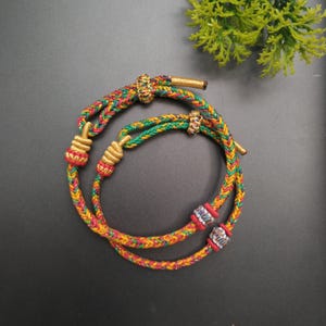 Pode incluir: Duas pulseiras de cordão trançado com uma mistura vibrante de fios vermelhos, verdes e amarelos. Cada pulseira é acentuada com contas cilíndricas de tom dourado metálico e contas prateadas texturizadas, finalizadas com pontas de cordão com nós.