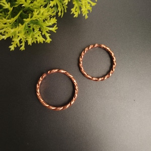 Puede incluir: Dos anillos circulares hechos de alambre de cobre con una textura trenzada similar a una cuerda. Cada anillo presenta una pequeña abertura, lo que indica un diseño abierto. La superficie metálica tiene un tono marrón rojizo cálido.