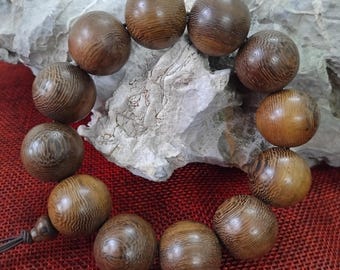 May include: A wooden bead bracelet featuring large, round beads with a natural wood grain pattern. The beads are strung together on a dark brown cord. The bracelet is displayed on a light gray stone surface against a red background.