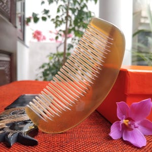 Puede incluir: Un peine curvo de color ámbar con dientes finos. El peine está hecho de un material translúcido y tiene un acabado liso y pulido. El peine descansa sobre una superficie roja, con una caja roja y una flor morada en el fondo.