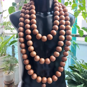 May include: A long, multi-layered necklace composed of smooth, round, light brown wooden beads with visible natural grain patterns. The beads are strung together in a continuous loop, draped in three distinct tiers over a black mannequin torso.