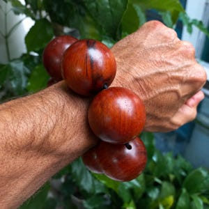 Peut inclure: Un bracelet composé de grosses perles en bois sphériques et lisses, présentant un grain brun rougeâtre profond. Les perles sont enfilées sur un cordon fin et sombre, avec une finition polie qui met en valeur la texture naturelle et les nuances du bois.