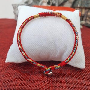 Può includere: Un braccialetto intrecciato rosso con un piccolo ciondolo a campana argentata. Il braccialetto presenta un design intrecciato con fili rossi, gialli, blu e dorati. Il braccialetto è esposto su un cuscino bianco su sfondo rosso.