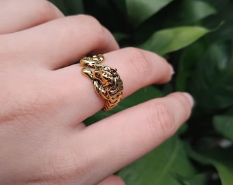 May include: A gold-colored ring featuring a detailed tiger design. The ring is wrapped around a finger, showcasing the intricate details of the tiger's face and body. The ring's design is a bold statement piece.