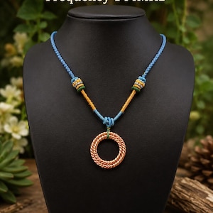 Può includere: Una collana con un ciondolo circolare in filo di rame intrecciato. Il cordino è azzurro con accenti decorativi gialli, verdi e blu. Il testo "Pure Copper Necklace Frequency 144MHz" è visualizzato sopra la collana.