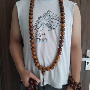 May include: A long necklace made of smooth, round, brown wooden beads worn around the neck, with a matching bracelet of similar dark brown wooden beads wrapped around the wrist. The beads are uniform in size and feature a polished, natural wood finish.