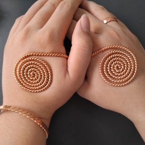 May include: A set of copper-colored jewelry featuring two spiral-shaped ornaments made of twisted rope-like metal, designed to rest on the back of the hands, accompanied by a matching twisted metal bracelet and a simple band ring on one finger.