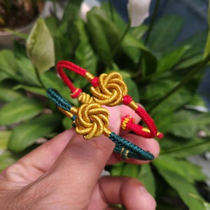 Puede incluir: Dos pulseras hechas a mano, una roja y dorada, la otra verde y dorada. Cada pulsera presenta un nudo decorativo en el centro. Las pulseras se exhiben sobre un fondo de follaje verde.